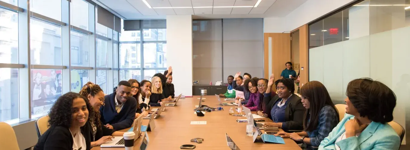 A look at a conference table from the head of the table. Over 16 people are talking among each other and themselves, with laptops in front of them, and three are raising their hands.
